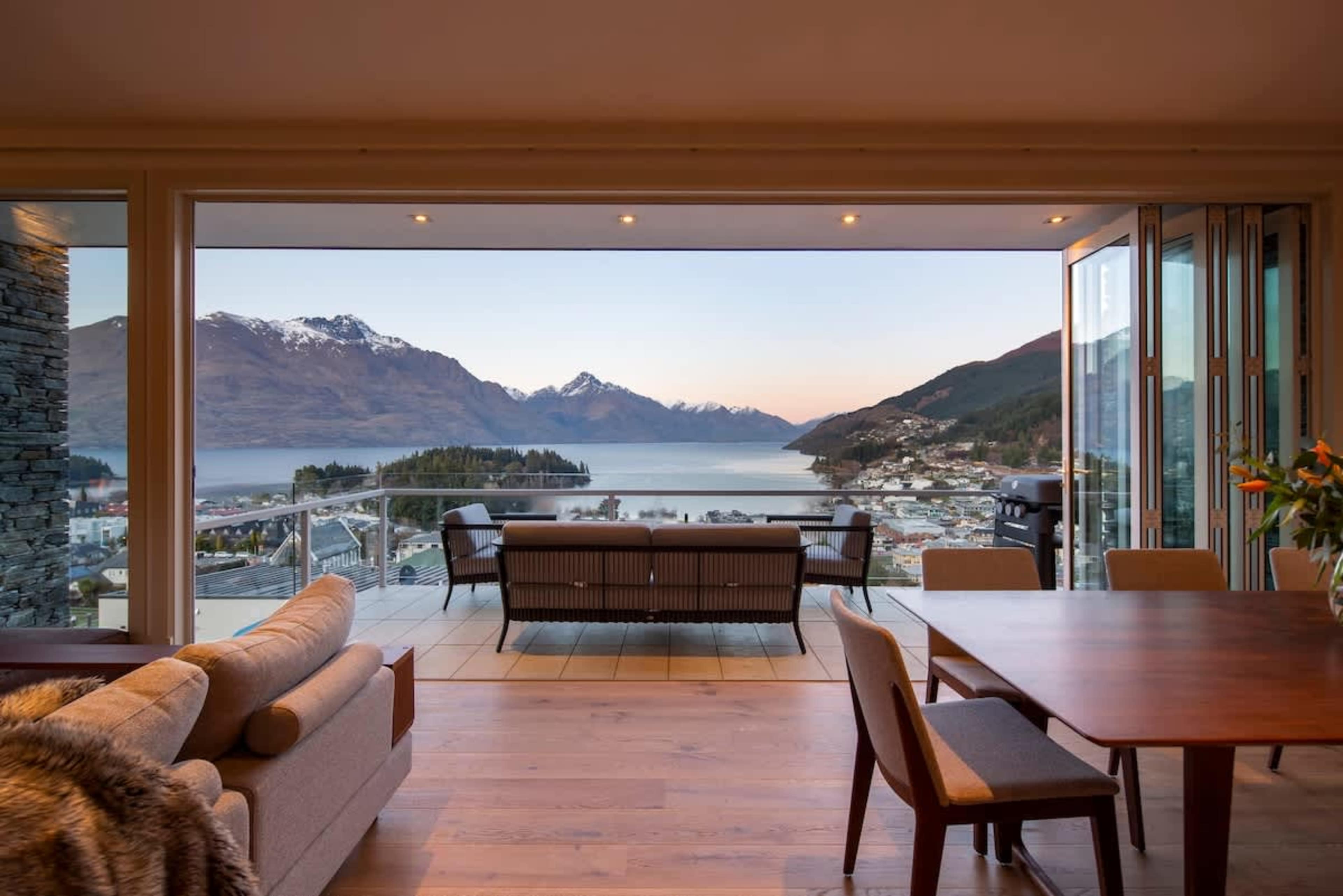 Living Room: Sun-drenched space with seating for five, gas fireplace, Smart TV, and sweeping lake and mountain views through floor-to-ceiling windows.