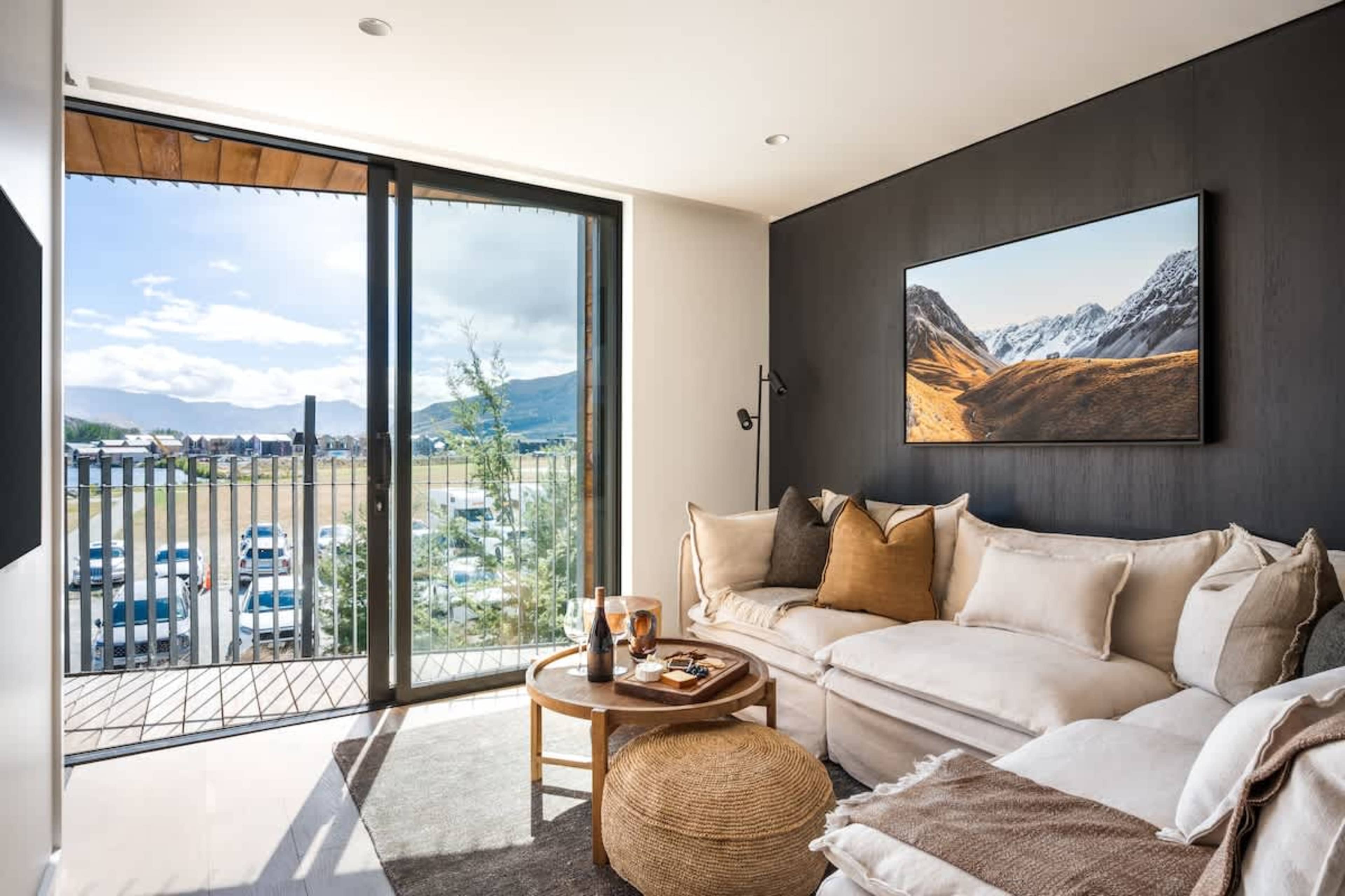 Luxury open-plan living room at Clubhouse Lane, Jacks Point Golf Course. Panoramic views of The Remarkables & Lake Tewa, modern alpine style, oak floors, vaulted cedar ceilings, fireplace, floor-to-ceiling windows. Queenstown golf & ski retreat.