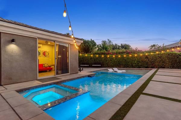 String lights reflect off pool and spa at dusk—casita glows warmly in the background beyond hedges.