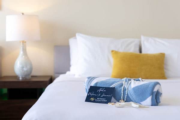 A neatly made bed with a yellow accent pillow and a welcome towel arrangement for a relaxing stay.