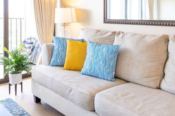 Bright living room with a beige sofa accented by blue and yellow pillows and natural light from the balcony.