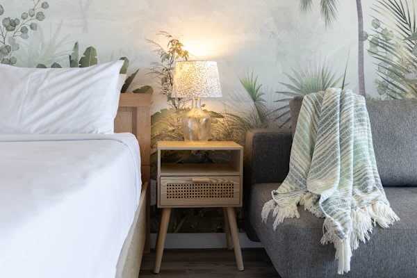 Details of the cozy queen-size bed, stylish nightstand featuring a charming lamp, and a futon sofa all set against a calming, tropical-inspired accent wall.