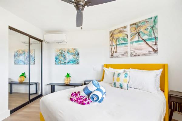 Unwind in this fresh, air-conditioned bedroom, offering a plush queen bed with a yellow headboard, full-length mirrored closets, and tropical island-themed artwork.
