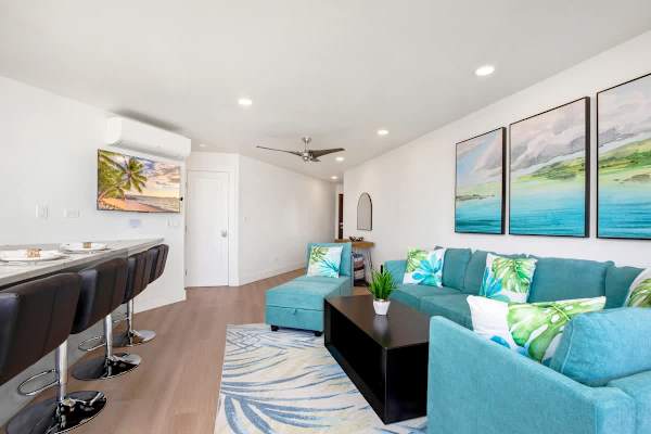 Relax in the bright, air-conditioned living area with a comfortable teal sectional sofa and stylish tropical decor, perfect for unwinding while watching the wall-mounted 50" Roku TV.
