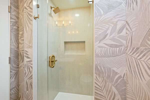 Step into this stunning walk-in shower featuring elegant tropical palm leaf accent tiling, a rain showerhead, and luxurious gold fixtures for a refreshing escape.