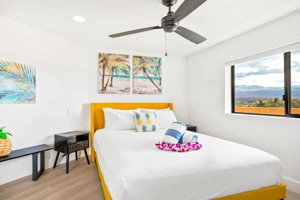 Retreat to this bright, primary bedroom featuring a comfortable queen bed with a vibrant yellow headboard, tropical artwork, and a large window offering great views.