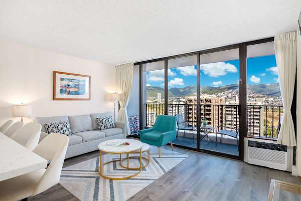 Bright living room with modern seating and a balcony overlooking city mountains.