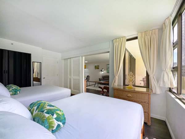 A bright bedroom featuring two queen beds, a wicker dresser, and sliding louvered doors that open to reveal the living area beyond, and a balcony view.