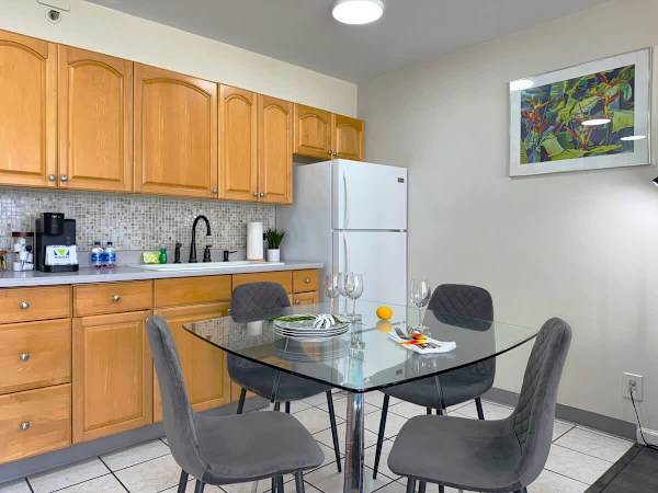 A bright kitchen and dining area featuring warm wood cabinets and a full-sized fridge, centered around a modern glass dining table set with plush, dark gray chairs.