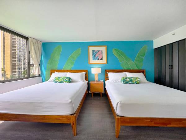 A stylish tropical bedroom featuring 2 comfy queen beds with matching wooden frames, centered on a vibrant teal accent wall painted with large banana leaves, and a bright city view through the window.