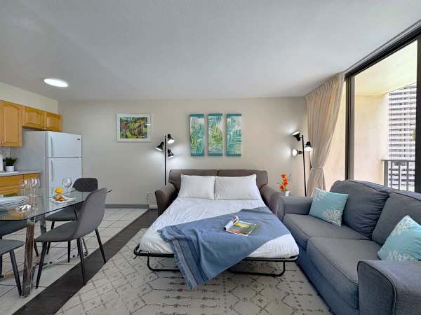 A versatile living area showcasing the sleeper sofa pulled out as an extra bed, set next to a cozy couch, a modern glass dining table, and a convenient full kitchen.