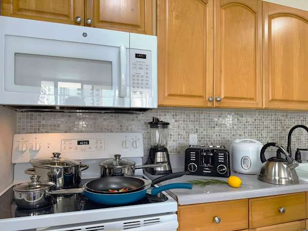 Well-stocked kitchen counter and stove with pots and pans, featuring light wood cabinets, a microwave, and several small appliances like a blender, toaster, and rice cooker.