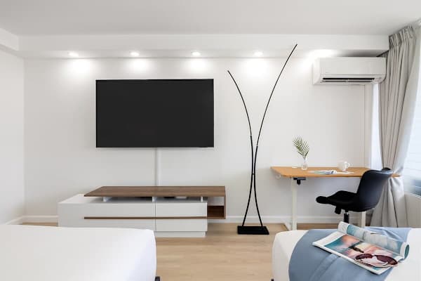 A sleek and bright studio apartment featuring a wall-mounted 65" smart TV, a stylish standing desk, split AC, and minimalist decor.