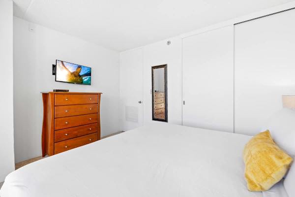 This clean, bright bedroom ensures entertainment with a mounted flat-screen TV above a spacious wooden dresser, complemented by large, convenient closet space.
