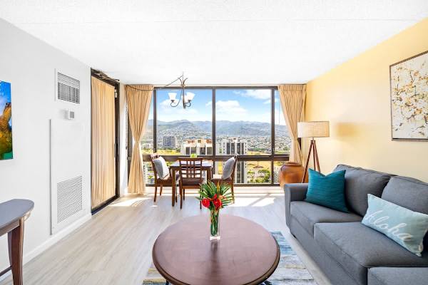 The living room features comfortable seating, a dining area, and stunning panoramic views of the city and mountains through large picture windows.