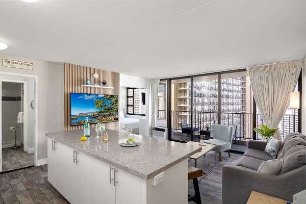 The living area features a kitchen island with seating, a cozy pull-out sleeper sofa, access to the balcony, and a stylish media center with a mounted 65" Smart TV.