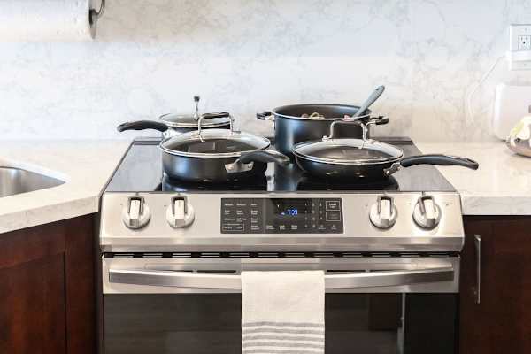 Fully equipped modern kitchen featuring a sleek glass cooktop stove/oven with multiple pots and pans ready for use, set against a marble-patterned backsplash.