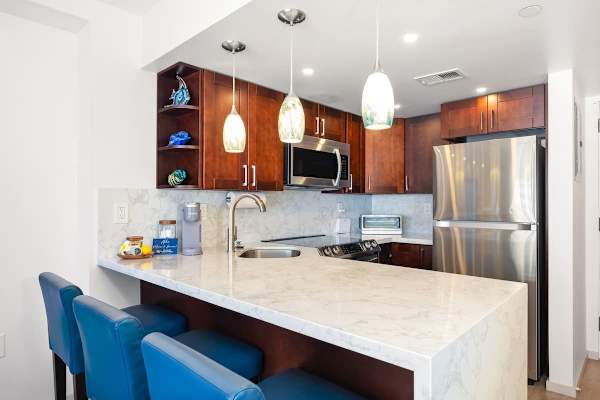 Your fully equipped kitchen features dark wood cabinetry, stainless steel appliances (including a refrigerator, microwave, and range), and a bright white marble countertop with an integrated sink and bar seating for three.