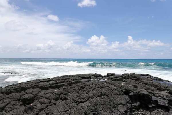Beautiful tropical coast view featuring rugged volcanic rock, a soft white sand beach, and crashing turquoise ocean waves under a sunny sky! 🌊🏖️☀️