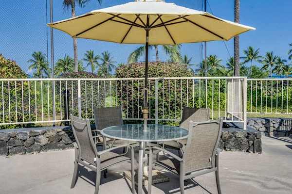 Patio dining set sits beneath umbrella with tropical greenery nearby