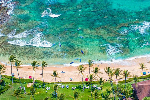Aerial view of the turquoise waters gently wash onto the white sandy beach at Kiahuna Plantation Resort, creating the perfect backdrop for a relaxing day by the ocean.
