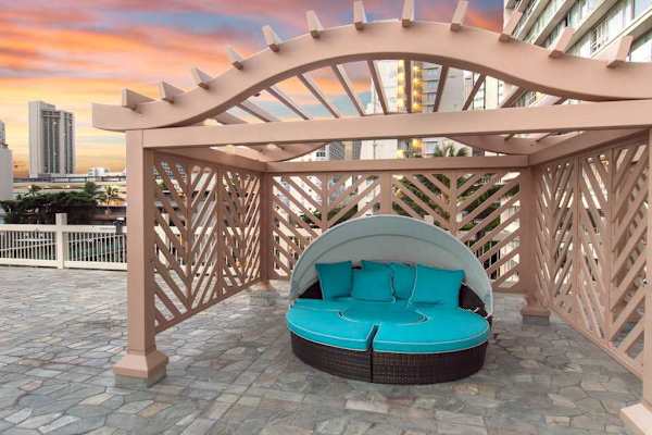 Lounge in ultimate comfort inside one of the shaded poolside cabanas, outfitted with a large round daybed and vibrant teal cushions under a decorative wooden pergola.