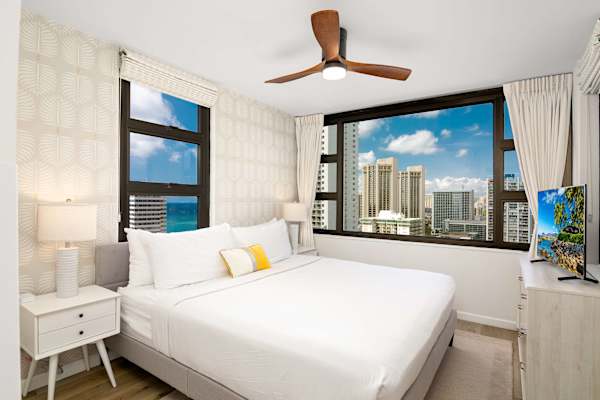 Bright bedroom w/ bed, ceiling fan & large windows showcasing city & ocean views