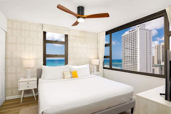 Sunlit bedroom w/ bed, ceiling fan & large windows framing ocean & city views