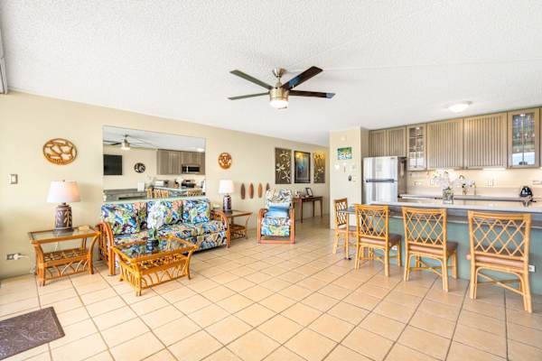 Relax in your bright and airy open-concept living area, showcasing a vibrant, tropical-printed sleeper sofa alongside a breakfast bar with wicker seating that transitions smoothly into the kitchen.