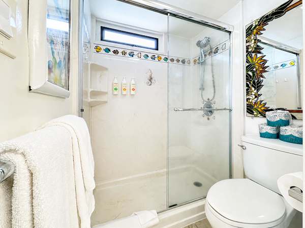 Unwind after a day at the beach in this clean, well-lit bathroom featuring a spacious walk-in shower with sliding glass doors, handy built-in shelving, and a bright tropical border