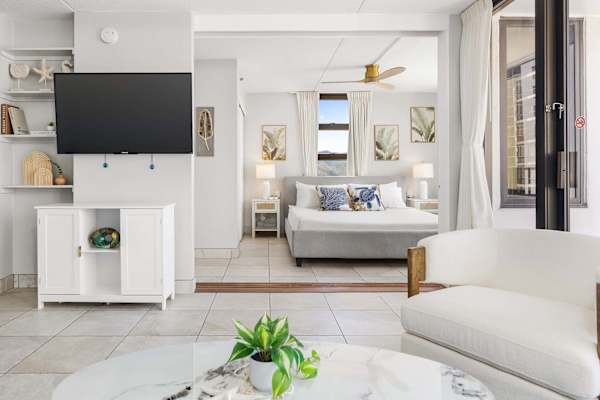 Your bright view shows the main living space with a wall-mounted 50" TV and white couch, looking directly into the bedroom area with a king-size bed, which is separated by a sliding door.