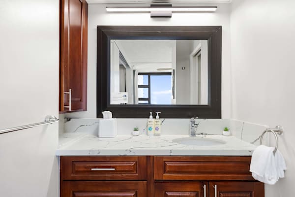 Your bathroom sink area features a white countertop, wooden cabinets, a sink with soap dispensers, a large dark-framed mirror, and a modern light above.