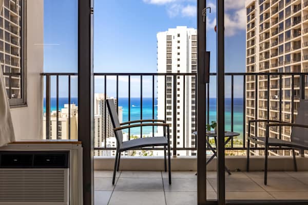 This view looks through the glass sliding door out to the balcony, showing the balcony chairs, a small table, an air conditioning unit inside, and the ocean and city skyline in the background.