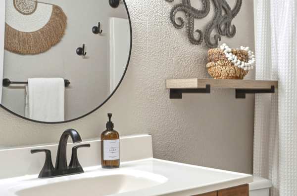 Your stylish bathroom vanity area features matte black fixtures, a round mirror, and chic coastal accents like woven wall art and decorative shelving.