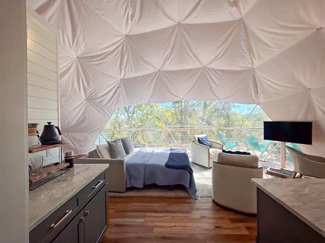 Bright dome interior with pull out bed, seating area, and large window framing wooded views.